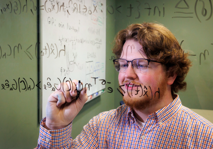 Male student writing math equations on the glass between him and the camera.